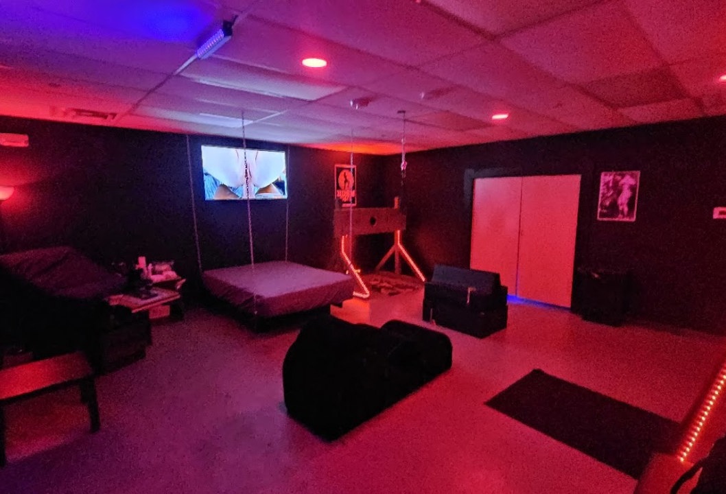 Red-lit BDSM playroom with suspended bed and lounge furniture
