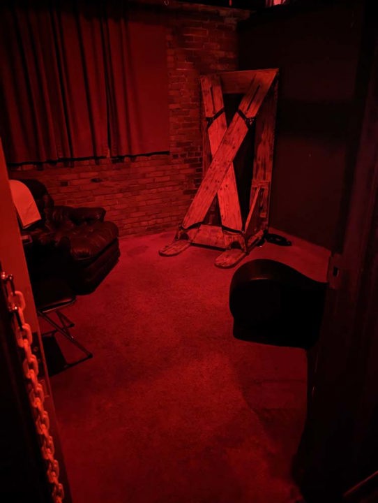 Red-lit themed play room with wooden restraint frame